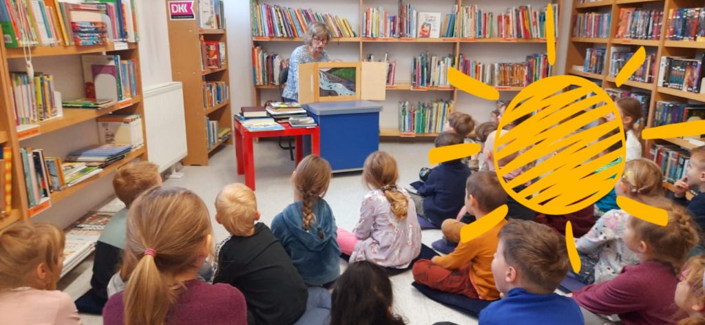 Zdjęcie przedstawia dzieci w bibliotece, siedzące na poduszkach, tyłem do aparatu, przodem do pani bibliotekarki, przedstawiającej dzieciom teatrzyk kamishibai. W tle półki z książkami, dwa stoliki - czerwony i granatowy, na granatowym pani ustawia drewniany teatrzyk. Po lewej, na czerwonym, leżą książki.