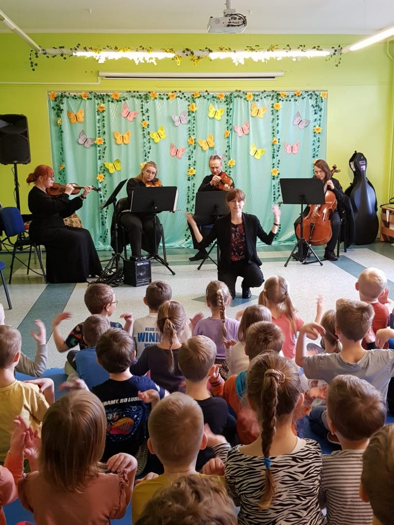 Zdjęcie przedstawia koncert muzyczny. Przed grupą dzieci kwartet smyczkowy oraz pani prowadząca koncert. Muzycy ubrani na czarno, siedzą na krzesłach przed pulpitami. W tle zielona zasłona ozdobiona kwiatami. Dzieci siedzą na podłodze w sali, wpatrzone w muzyków, tyłem do obiektywu aparatu. Pani prowadząca koncert zwrócona jest przodem do dzieci, tyłem do muzyków, kuca przed dziećmi unosząc obie ręce.