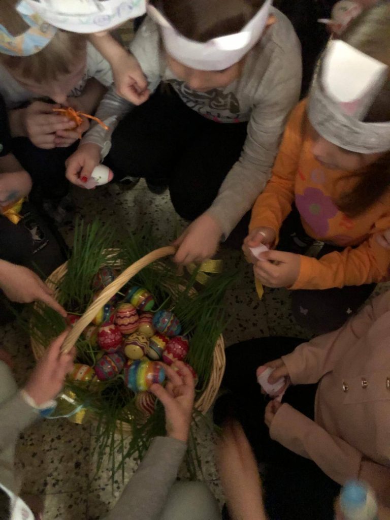 Wokół koszyka z kolorowymi jajami zgromadziło się kilkoro dzieci, które sięgają dłońmi do koszyka, aby wyjąć jaja.