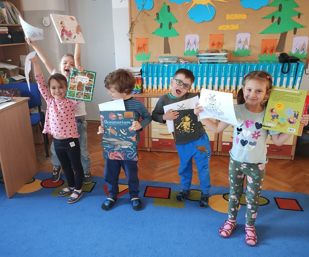 Pięcioro dzieci stoi w sali przedszkolnej, trzymając w rękach uniesione książki oraz kartki z wizerunkiem mola książkowego. Dzieci stoją na niebieskim dywanie. W tle tablica korkowa z papierowymi ozdobami i napisami: 