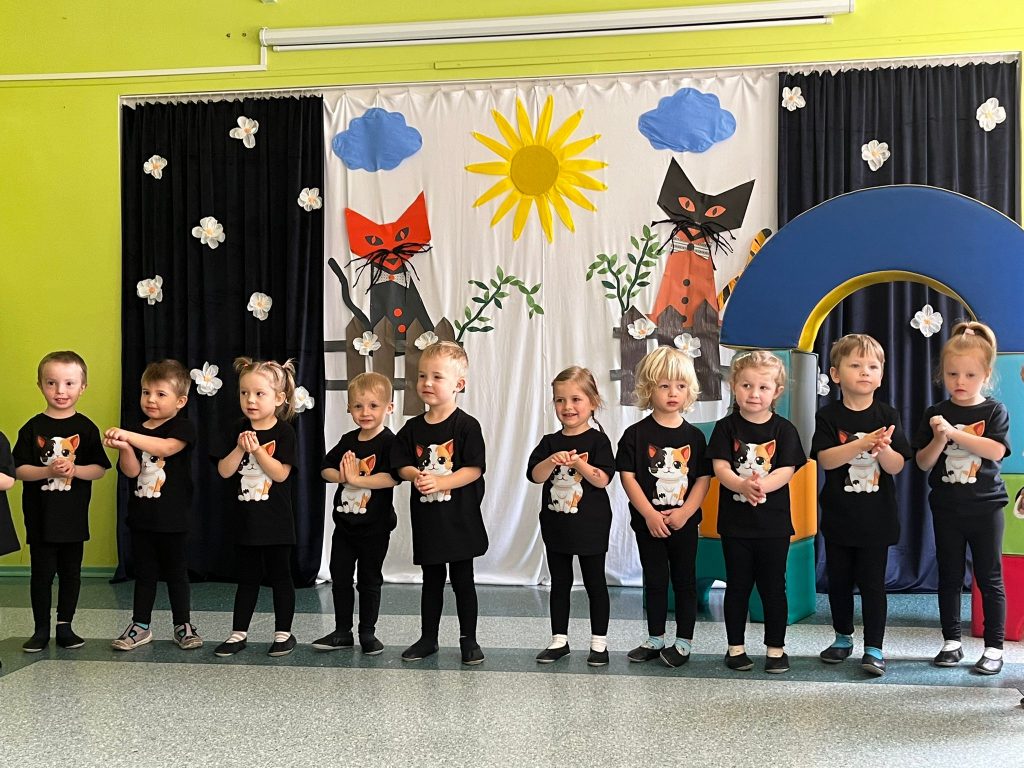 Dzieci ubrane w czarne stroje z wizerunkami kotków na koszulkach, śpiewają, stojąc z uniesionymi na wysokość pasa rączkami. Dzieci stoją w rzędzie, na ciemnozielonym pasie. W tle dekoracja przedstawiająca dwa siedzące na płotach, czarno-pomarańczowe koty.