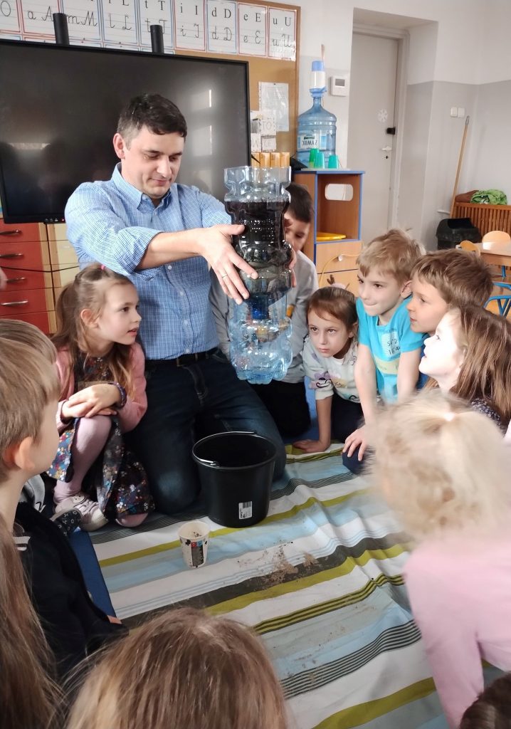 Mężczyzna w niebieskiej koszuli trzyma dłońmi nałożone na siebie dwie plastikowe butle, wypełnione węglem, ziemią, piaskiem, tworząc filtr, przez który przelewa się woda. Wokół siedzą dzieci, wpatrujące się w przepływającą wodę. W tle czarny ekran, nad nim plansze z literami. Obok butla z wodą pitną i jasne drzwi do sali. Dzieci siedzą z otwartymi szeroko oczami, wokół pasiastej ceraty.