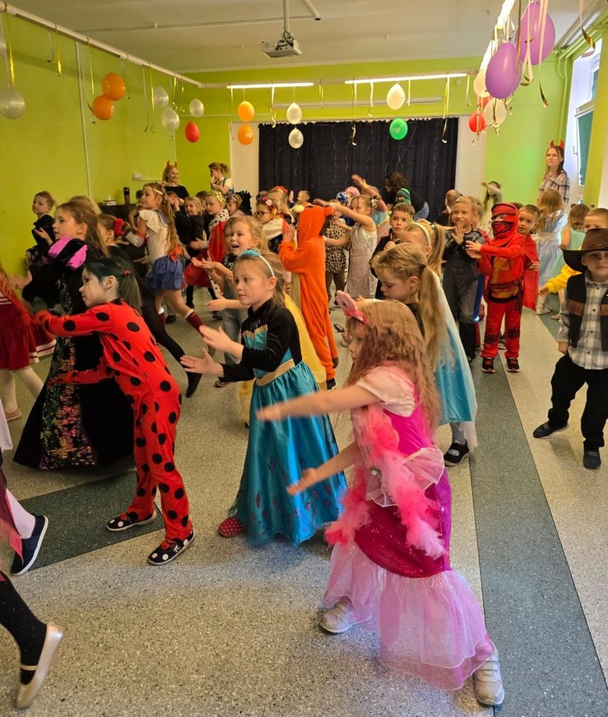 Zabawa karnawałowa w przedszkolu. Duża grupa kolorowo przebranych dzieci tańczy w zielonej sali, kierując ręce w swoją lewą stronę. Na pierwszy planie dziecko w czerwonym kombinezonie w czarne kropki, pośrodku dziewczynka w długiej turkusowej, błyszczącej sukni z czarnymi rękawami, z przodku dziewczynka w różowej sukni. U sufitu zawieszone są kolorowe balony.