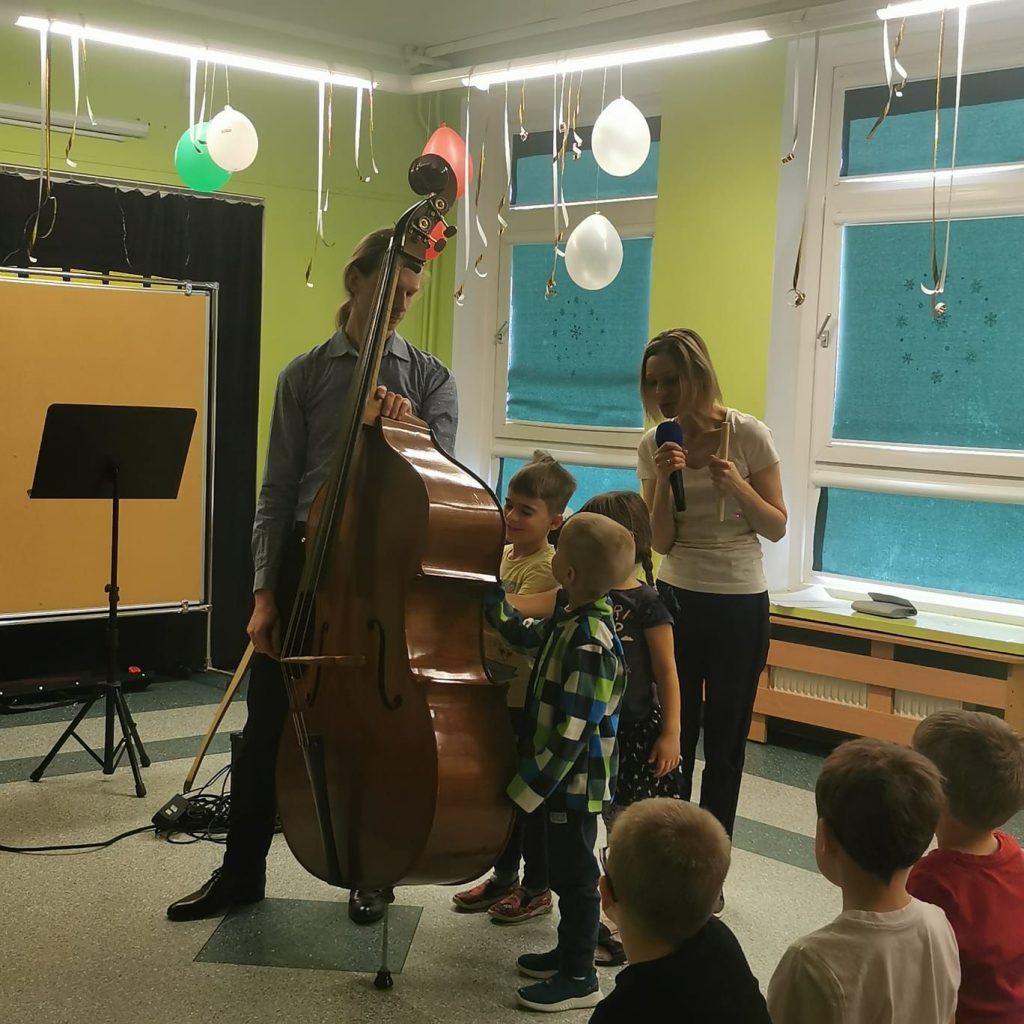 Koncert dla dzieci. Na środku kontrabasista stojący z kontrabasem. Obok troje dzieci, które z bliska spoglądają na instrument, jedno dziecko dotyka go. Za nimi pani w jasnej bluzce, z mikrofonem w prawej ręce. Pochyla się nad dziećmi i coś do nich mówi. W tle zielone rolety na oknach z białymi ramami. Balony zwisające u sufitu. Po lewej fragment tablicy korkowej w kolorze beżowym, a przed nią czarny pulpit.