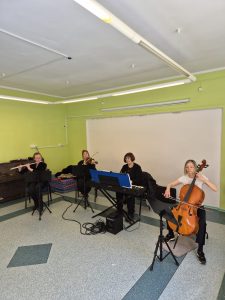 Na zdjęciu kwartet muzyków. Od lewej: flecistka, skrzypaczka, pianistka przy pianinie elektryczym, wiolonczelistka. Panie z instrumentami siedzą na krzesłach. W tle jasna, kremowa wnęka …<p class=