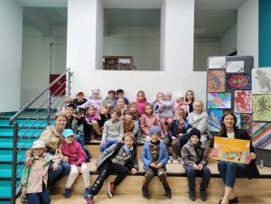 Duża grupa dzieci wraz z trzema osobami dorosłymi. Wszyscy siedzą na schodach w holu szkoły. W tle białe ściany z wyciętymi kwadratowymi wnękami. Na dolnym schodku, po prawej stronie, siedzi pani wicedyrektor szkoły, trzymająca przed sobą żółtą kartkę z napisem "Wesołych Świąt" i kolorowymi papierowymi jajami. Po lewej stronie poręcz schodów i fragment schodów w kolorze zielonym. Po prawej, za panią wicedyrektor, tablica z pracami dzieci.