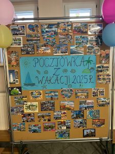 Na korkowej, beżowej tablicy, znajdują się liczne pocztówki dzieci z wakacji (znad morza, z gór, jezior oraz wyjazdów zagranicznych), opatrzone licznymi obrazkami i życzeniami. Na środku umieszczony jest niebieski brystol z napisem ,,Pocztówka z wakacji 2025 r". Wokół napisu narysowane są również: słońce, żaglówka, muszelki i palma. Tablicę przystrajają 4 kolorowe balony.