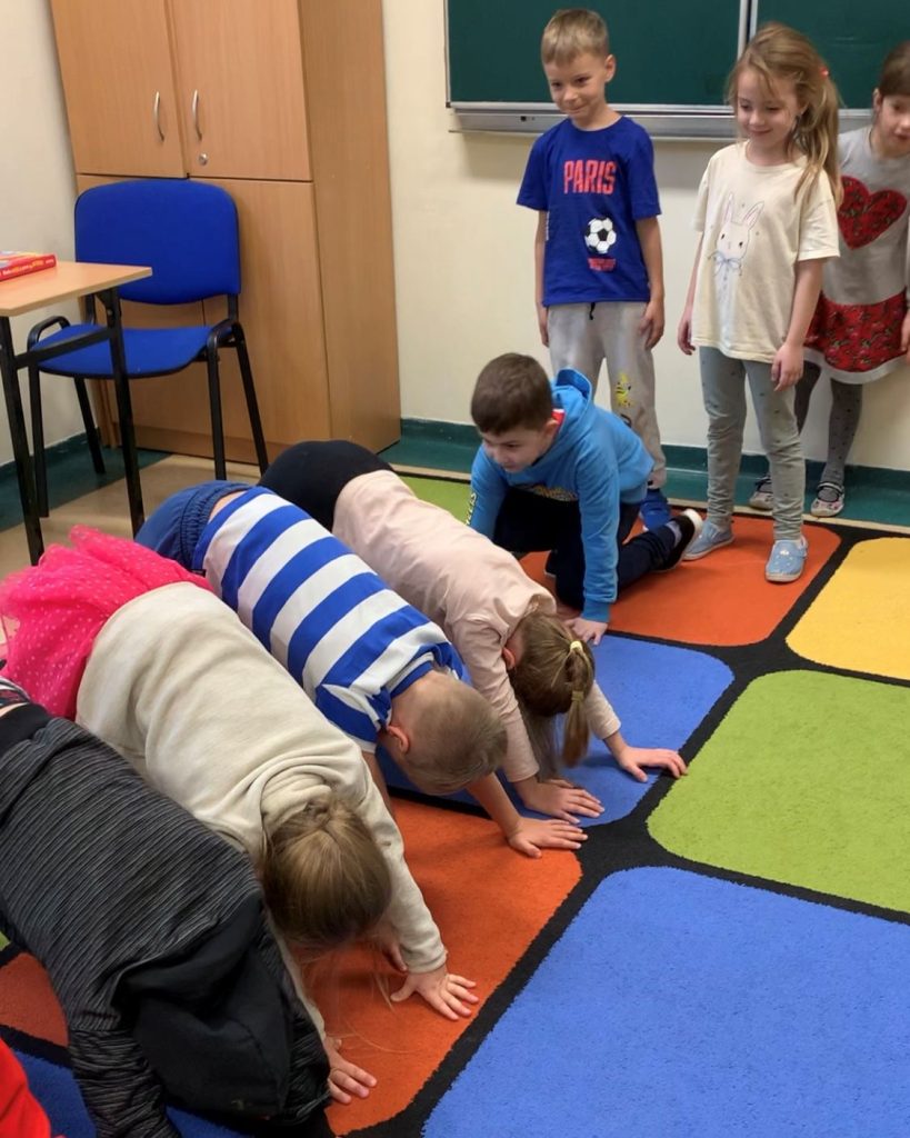 Zajęcia sportowe w przedszkolu. Kilkoro dzieci wykonuje skłon z oparciem dłoni o dywan, tworząc tunel, pod którym mają przejść inne dzieci. Przed nimi ustawiają się dzieci, które będą przez tunel przechodzić. Dzieci ćwiczą na kolorowym dywanie w kwadraty. W tle po lewej stronie małe biurko i niebieskie krzesło. Za nim wysoka beżowa szafa. Za dziećmi stojącymi zielona tablica.