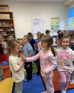 Grupa dzieci bierze udział w zajęciach ruchowych. Dwie dziewczynki dobrane w pary, zwrócone buziami do siebie, dotykają się dłońmi. Po prawej stronie dziewczynka w jasnej bluzie z różowym napisem i rysunkiem, zwrócona buzią do obiektywu aparatu. Za parą dziewczynek i tą dziewczynką pozostałe dzieci, poszukujące par. W tle jasna ściana z zawieszonymi dwoma afiszami. Po lewej stronie regał z książkami.