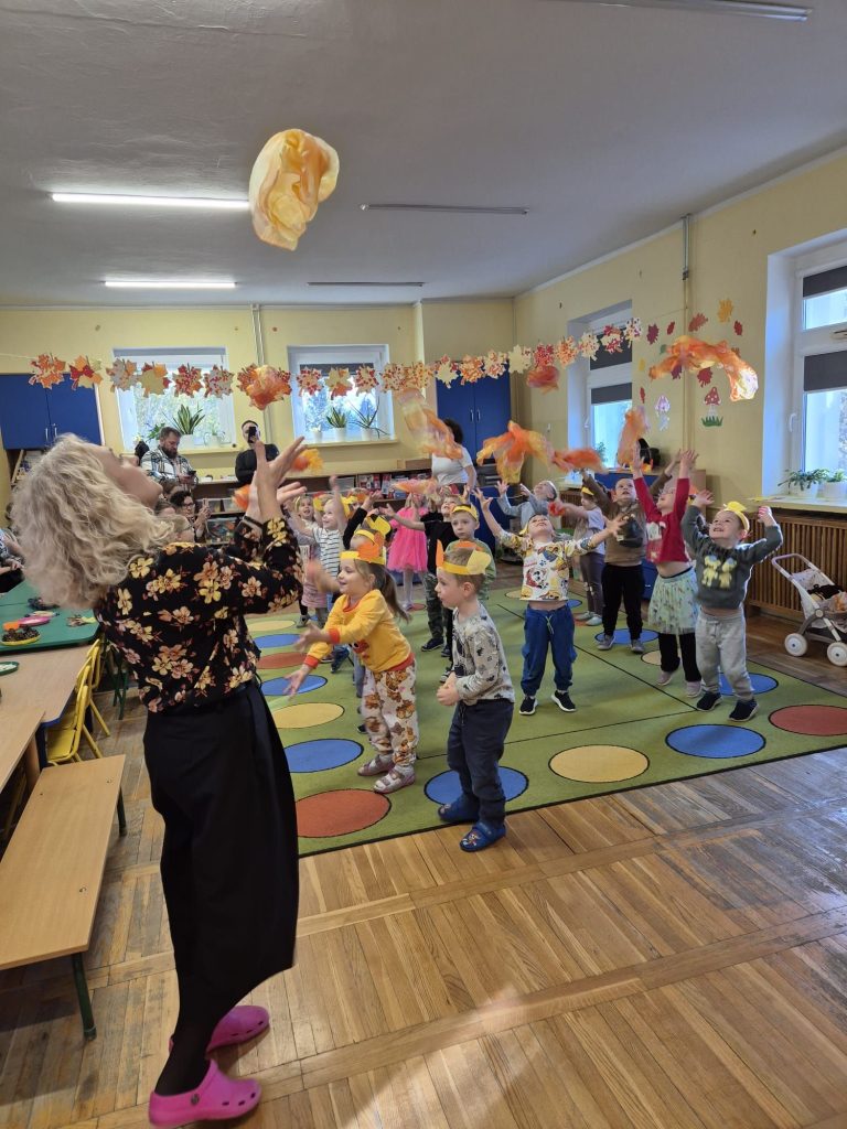Nauczycielka wraz z grupą dzieci poruszają się po drewnianej podłodze oraz zielonym dywanie w kolorowe, duże koła. Dzieci mają na sobie opaski z papierowymi liśćmi. Nauczycielka i niektóre dzieci podrzucają jasny worek. W tle podłużne lampy na suficie, sznurek z pracami dzieci zawieszony pod sufitem.