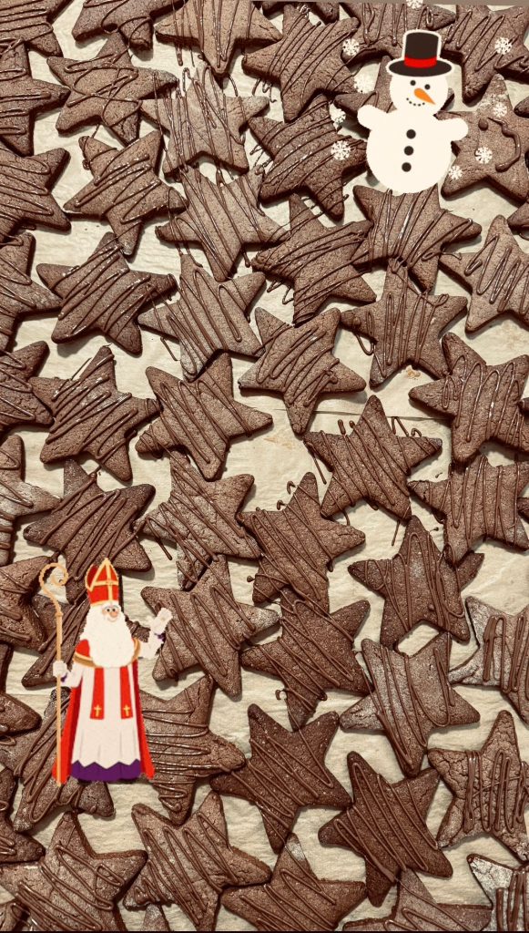 Brązowe, czekoladowe ciastka-gwiazdki, ozdobione brązowymi, ukośnymi paskami czekolady, rozłożone ba blacie. W lewym dolnym rogu postać Świętego Mikołaja, biskupa. W prawym górnym rogu postać białego bałwanka w czarnym kapeluszu z czerwoną wstążką, pomarańczowym nosem z marchewki, trzema czarnymi guzikami.