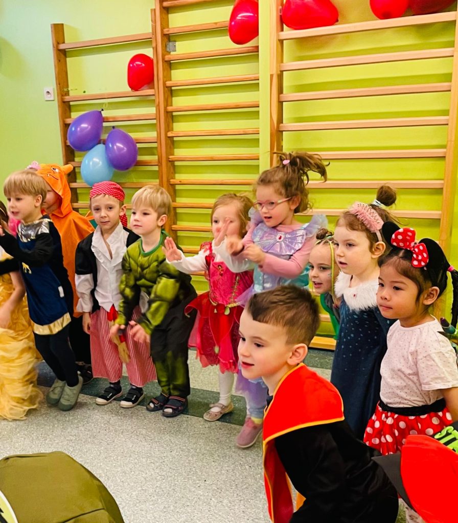 Grupa dzieci w strojach karnawałowych stoi tyłem do drabinek gimnastycznych. W tle jasnozielona ściana, kilka balonów - dwa fioletowe, jeden błękitny, dwa czerwone. Dzieci delikatnie kucają lub podskakują, patrząc w jednym kierunku.