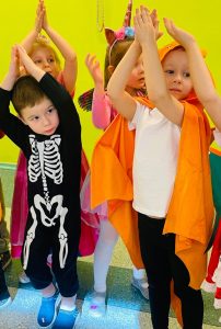 Czworo dzieci w strojach karnawałowych, z uniesionymi rękami. Z przodu po lewej chłopiec w stroju czarnym z biały szkieletem. Obok chłopiec w białym T-shircie i czarnych spodniach, w pomarańczowej pelerynie. Za nimi dziewczynki.