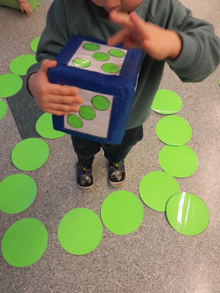 dziecko trzyma kostkę z dużymi, zielonymi oczkami. Stoi na szarej podłodze. Wokół niego duże zielone koła, leżące na podłodze.