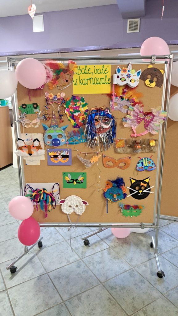 Korkowa tablica w korytarzu przedszkola z kolorowymi, wykonanymi prze dzieci i rodziców z papieru maskami karnawałowymi. U góry napis na żółtym tle: 