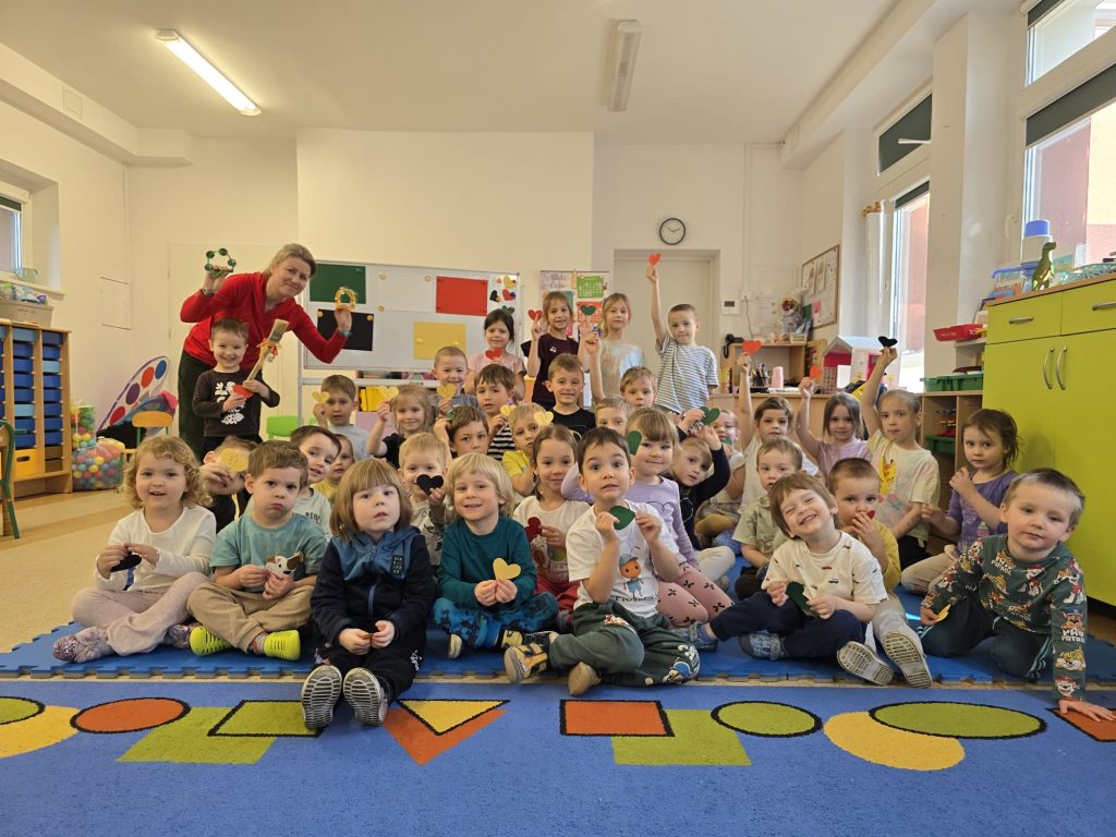 Grupa dzieci wraz z nauczycielką po lewej stronie, siedzi na dywanie. Dzieci z ostatniego rzędu stoją. Dzieci trzymają w dłoniach papierowe serduszka. W tle biała tablica z kolorowymi arkuszami papieru. Dzieci siedzą na niebieskim dywanie w pomarańczowe, żółte i zielone figury geometryczne. Nauczycielka trzyma w dłoniach małe tamburyna. Ubrana jest czerwoną bluzkę.