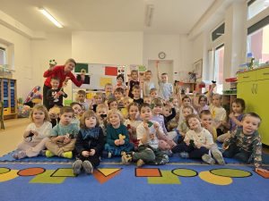 Grupa dzieci wraz z nauczycielką po lewej stronie, siedzi na dywanie. Dzieci z ostatniego rzędu stoją. Dzieci trzymają w dłoniach papierowe serduszka. W tle biała tablica z kolorowymi arkuszami papieru. Dzieci siedzą na niebieskim dywanie w pomarańczowe, żółte i zielone figury geometryczne. Nauczycielka trzyma w dłoniach małe tamburyna. Ubrana jest czerwoną bluzkę.