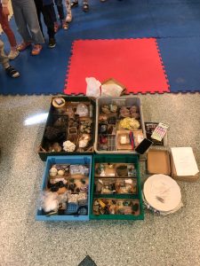 Cztery duże płaskie, prostokątne pojemniki z minerałami w formie kamieni. Obok mniejsze pojemniki z minerałami w formie proszku. Za pojemnikami czerwono-granatowa mata. Po lewej stronie widać nogi dzieci w obuwiu.