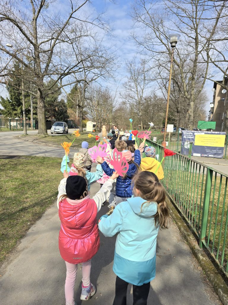 Grupa dzieci idzie wzdłuż zielonego ogrodzenia, niosąc ze sobą papierowe kwiatki w ręku.