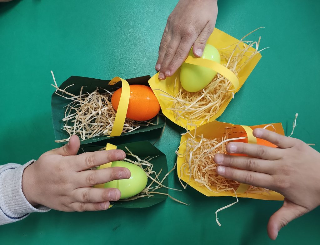 Na zielonym stole cztery papierowe koszyczki z żółtym papierowym paskiem. W koszyczku słoma, na niej plastikowe jaja: dwa pomarańczowe, dwa żółte. Dłonie dzieci nad trzema koszyczkami.