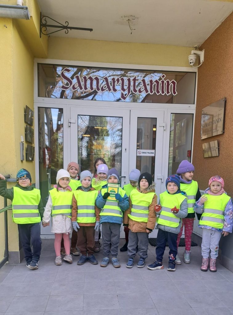 Grupa dzieci w żółtych odblaskowych kamizelkach stoi przed drzwiami budynku Domu Pomocy Społecznej. Wysoko na drzwiach napis 
