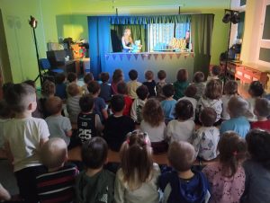 Grupa zwrócona w kierunku niebieskiej kotary, odsłaniającej scenkę odgrywanego dla nich przedstawienia teatralnego. Kolor kotary jest niebieski. W jej okienku aktorka trzymająca pacynkę - niebieskiego psa. W tle zielona ściana.